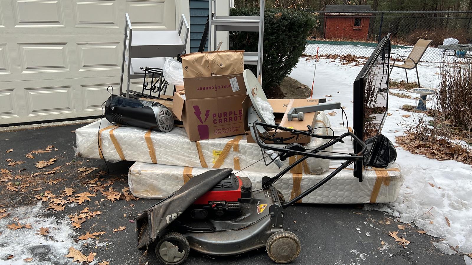 An old lawnmower, two mattresses, a stereo, a television, and some other junk sitting in a driveway outside of a garage